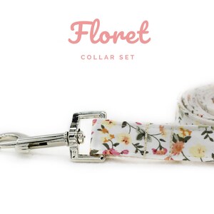 K&ouml;nnte beinhalten: Ein wei&szlig;es Hunde-Halsband- und Leinen-Set mit Blumenmuster und einem silbernen Metallverschluss und Haken. Leine und Halsband zeigen ein Muster aus kleinen gelben, rosa und orangefarbenen Blumen. Der Text "Floret COLLAR SET" ist in Rosa geschrieben.
