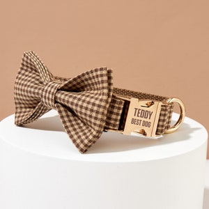 May include: A brown and tan checked dog collar with a gold buckle. The buckle has the text "Teddy Best Dog" engraved on it. The collar has a matching brown and tan checked bow tie attached.