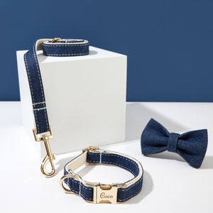 May include: A blue denim dog collar and lead set with a gold buckle and clasp. The collar has the word "Coco" engraved on the buckle. The set is displayed on a white surface with a blue denim bow tie.