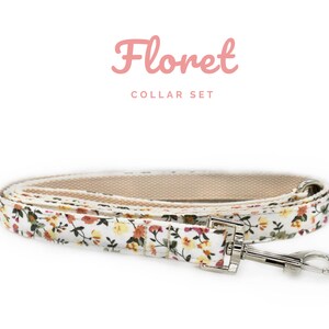K&ouml;nnte beinhalten: Ein floral bedrucktes Hunde-Leinen- und Halsband-Set. Die Leine und das Halsband haben einen wei&szlig;en Hintergrund mit einem Muster aus kleinen gelben, orangefarbenen und gr&uuml;nen Blumen. Die Leine hat einen silbernen Metallverschluss und eine beige Innenseite.