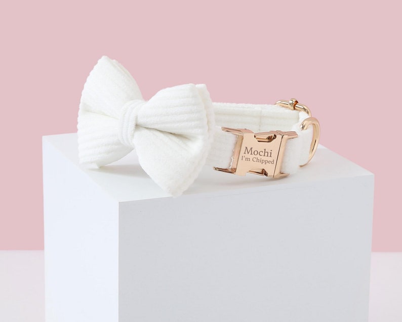 May include: A white corduroy dog collar and matching bow tie set. The collar features a rose gold buckle engraved with "Mochi I'm Chipped." The bow tie is a simple, elegant design. The set is displayed on a white cube against a pink background.