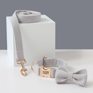 May include: A light gray corduroy dog collar, leash, and bow tie set. The collar has a gold-colored buckle with the name "TEEDY" engraved. The leash has a matching gold-colored clasp. The bow tie is a simple, pre-tied design.