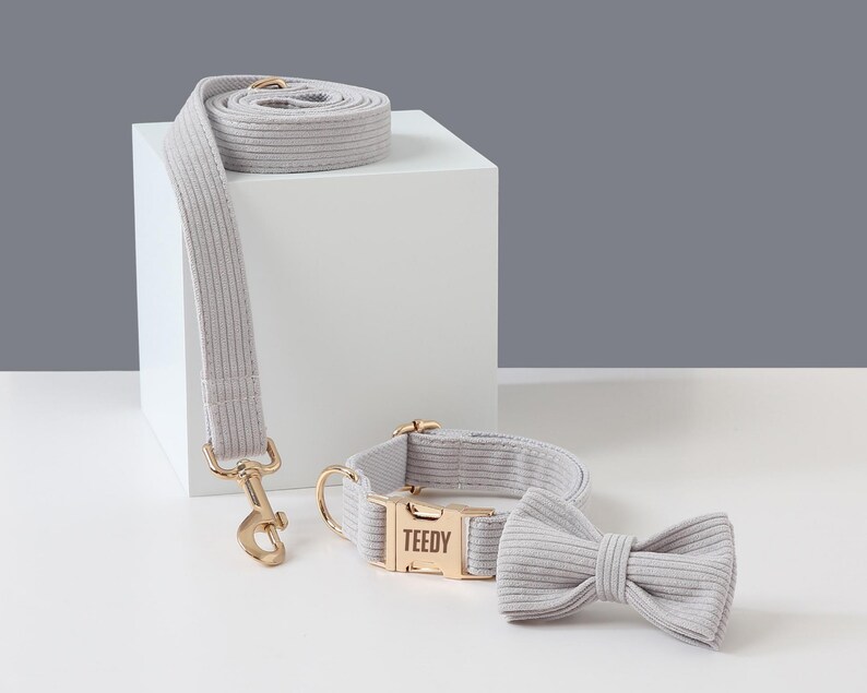 May include: A light gray corduroy dog collar, leash, and bow tie set. The collar has a gold-colored buckle with the name "TEEDY" engraved. The leash has a matching gold-colored clasp. The bow tie is a simple, pre-tied design.
