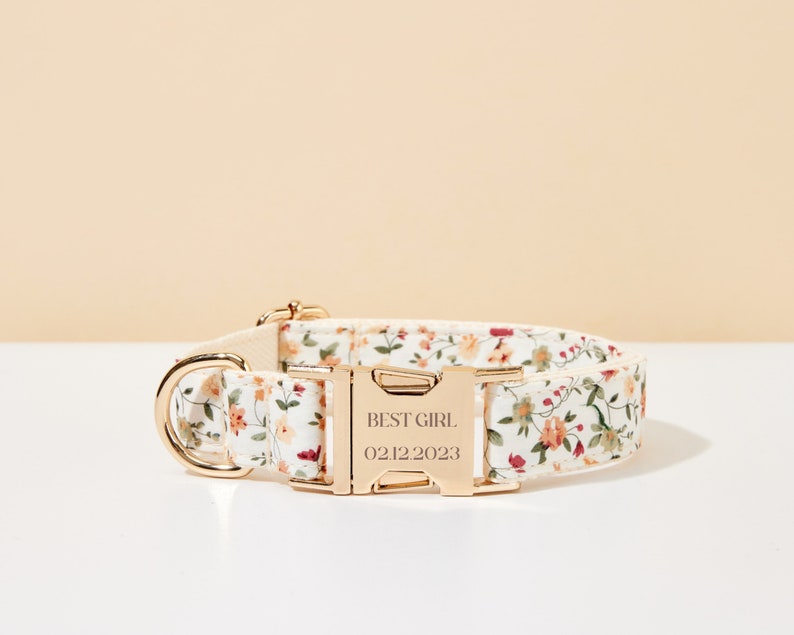 K&ouml;nnte beinhalten: Ein Hundehalsband mit floralem Muster und goldfarbenen Metallbeschl&auml;gen. Das Halsband hat einen wei&szlig;en Hintergrund mit einem Muster aus kleinen orangefarbenen, rosa und gr&uuml;nen Blumen. Die Schnalle ist mit "BEST GIRL" und dem Datum 02.12.2023 graviert.