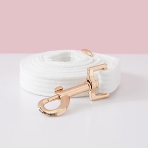 May include: A white dog leash with a gold-colored metal clasp and hardware is coiled on a white surface. The leash is made of a soft, textured material. The background is a light pink.