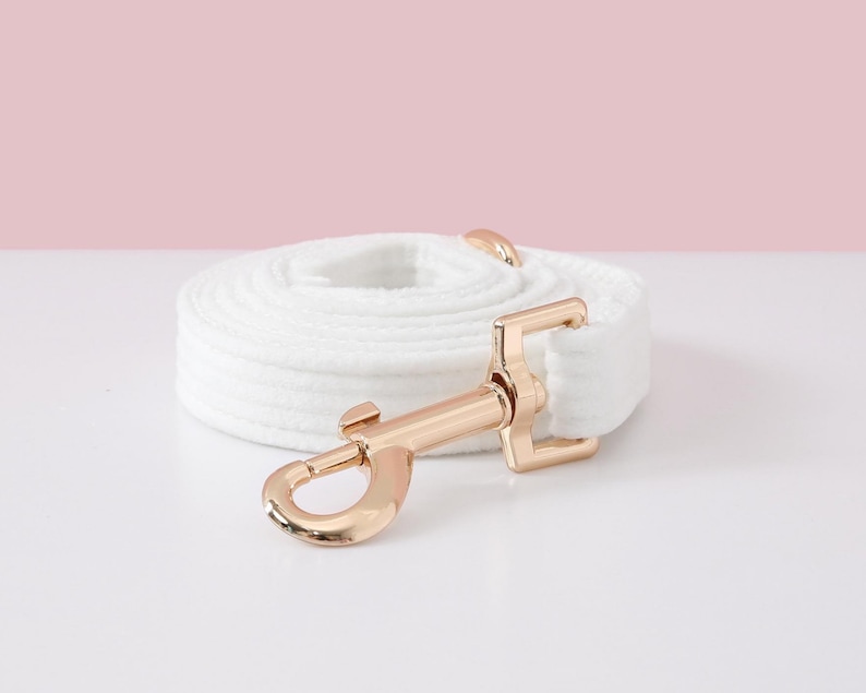May include: A white dog leash with a gold-colored metal clasp and hardware is coiled on a white surface. The leash is made of a soft, textured material. The background is a light pink.
