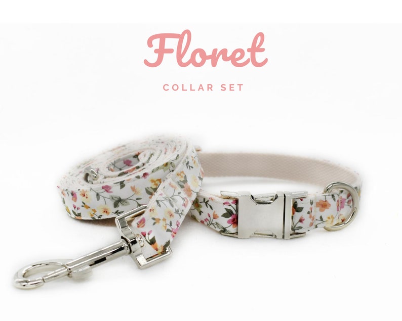 K&ouml;nnte beinhalten: Ein floraler Halsband-Set mit einem wei&szlig;en Halsband und einer passenden Leine, verziert mit einem Muster aus kleinen rosa, gelben und orangefarbenen Blumen. Das Set enth&auml;lt eine silberfarbene Metallschnalle und einen Leinenverschluss. Der Text "Floret COLLAR SET" ist rosa.