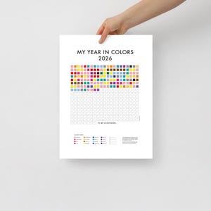 May include: A white poster titled "MY YEAR IN COLORS 2026" with a grid of colorful squares. The top section is filled with colored squares, and the bottom section is blank. A color chart is at the bottom. The poster is held by a person.