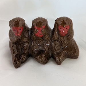 May include: Three brown ceramic monkeys, each with red painted faces, are sitting side-by-side. The monkey on the left has its hands covering its ears, the middle monkey has its hands covering its eyes, and the monkey on the right has its hands covering its mouth.
