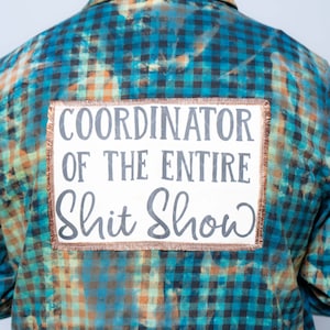 Może przedstawiać: Niebiesko-czarno-pomarańczowa koszula w kratę z białą naszywką. Na naszywce szarymi literami napisano "COORDINATOR OF THE ENTIRE Shit Show" i ma miedzianą obwódkę.