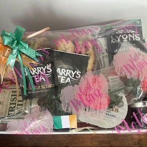 May include: A gift basket filled with Irish tea, cookies, and other treats, wrapped in clear cellophane and tied with a green and gold bow. The packaging includes text that says "Barry's Tea" and "Lyons Irish Cream". A small Irish flag is also visible.