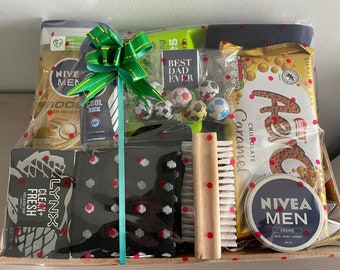 May include: A gift basket filled with various men's grooming products, including body wash, deodorant, soap, and hand cream. The basket also includes a chocolate bar, a brush, and a pair of socks. The basket is decorated with a green bow and red polka dots.