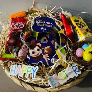 Puede incluir: Una cesta de mimbre llena de golosinas de Pascua, que incluye barras Kit Kat, Dairy Milk Buttons y Mini Eggs. La cesta está decorada con hierba decorativa, una zanahoria y un letrero de "Happy Easter".