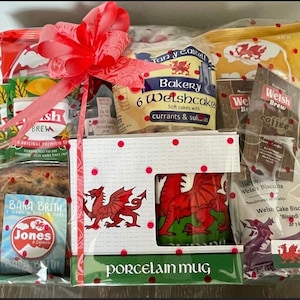 May include: A gift basket with a red bow, featuring Welsh cakes, tea, coffee, biscuits, and a porcelain mug with a red dragon design. The basket is wrapped in clear cellophane and has a red and white polka dot pattern.
