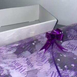 May include: A white gift box with a purple ribbon and bow. The box is wrapped in clear plastic with a purple and white patterned design.