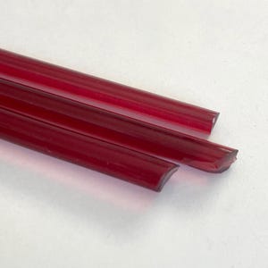 Pode incluir: Três varetas de vidro vermelho rubi translúcido dispostas diagonalmente sobre um fundo branco. As varetas têm formato retangular com bordas arredondadas. As extremidades são cortadas em ângulo, revelando uma superfície irregular.
