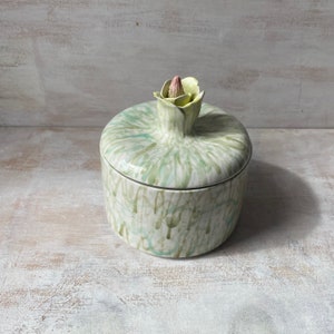 Puede incluir: Una caja de cerámica para guardar pequeños objetos con un patrón abstracto verde y blanco. La tapa tiene una flor delicada con un centro rosa y pétalos verdes.