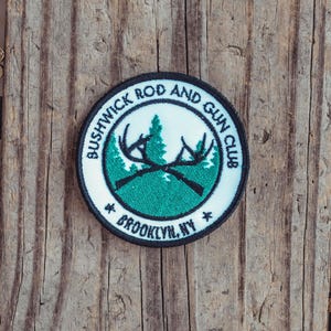 May include: A black and white embroidered patch with a green background. The patch features a crossed rifle and antlers with the text "Bushwick Rod and Gun Club Brooklyn, NY".