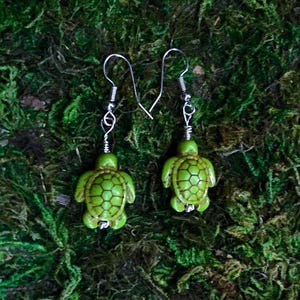 May include: A pair of green turtle earrings with silver-colored hooks. The turtle pendants have a textured shell design and are suspended from coiled wire. The earrings are displayed on a bed of green moss.