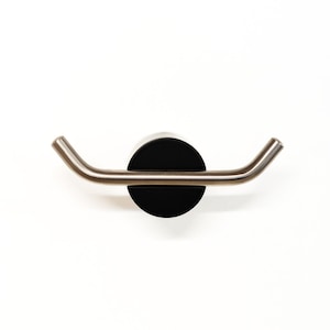 May include: A contemporary wall-mounted coat hook with a black circular base and a brushed metal bar. The bar curves outwards at each end to form two hooks. The design is minimalist and suitable for a modern interior. The dimensions are approximately 15 cm wide.