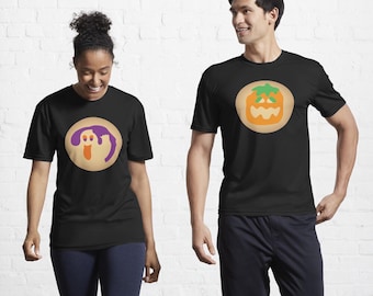 May include: Two black short-sleeved t-shirts. The left shirt features a circular graphic of a purple and orange ghost. The right shirt features a circular graphic of an orange and green jack-o'-lantern. Both shirts have a crew neckline.