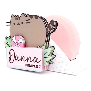 May include: A whimsical birthday card featuring a cartoon cat, a pink and white swirled lollipop, and the name "Danna" with "Cumple 7" printed on a pink card. The card has a pink balloon and mint green leaves.