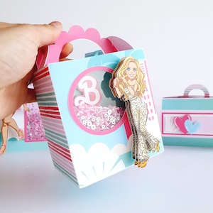 May include: A decorative gift box with a pink scalloped handle and a clear window filled with pink beads. The box features a cartoon illustration of a woman in a gold jumpsuit. The box has a light blue background with pink and green stripes.