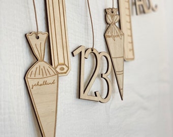 May include: Wooden ornaments with school-themed designs, including a pencil, a ruler, a cone with the word "schulkind" written on it, and the numbers 123. The ornaments are hanging from strings.