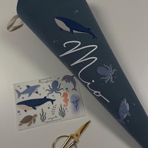 Könnte beinhalten: Ein dunkelblauer Stoffkegel mit Wal- und Oktopus-Motiv sowie dem Namen Mio in weißer Schreibschrift. Daneben liegt ein Bogen mit Bügelbildern BÜGELBILD SET Meerestiere und eine Schere aus goldfarbenem Metall.