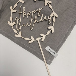 Könnte beinhalten: Ein Holz-Kuchentopper mit einem Kranz-Design und den Worten "Happy Birthday" in Kursivschrift. Der Topper befindet sich auf einem grauen Stoffhintergrund.