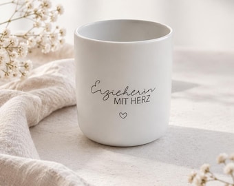 May include: A matte white ceramic mug featuring the black printed text "Erzieherin MIT HERZ" and a small heart icon centered below the lettering. The mug has a simple, handleless cylindrical shape with a smooth, clean finish and a flat base.
