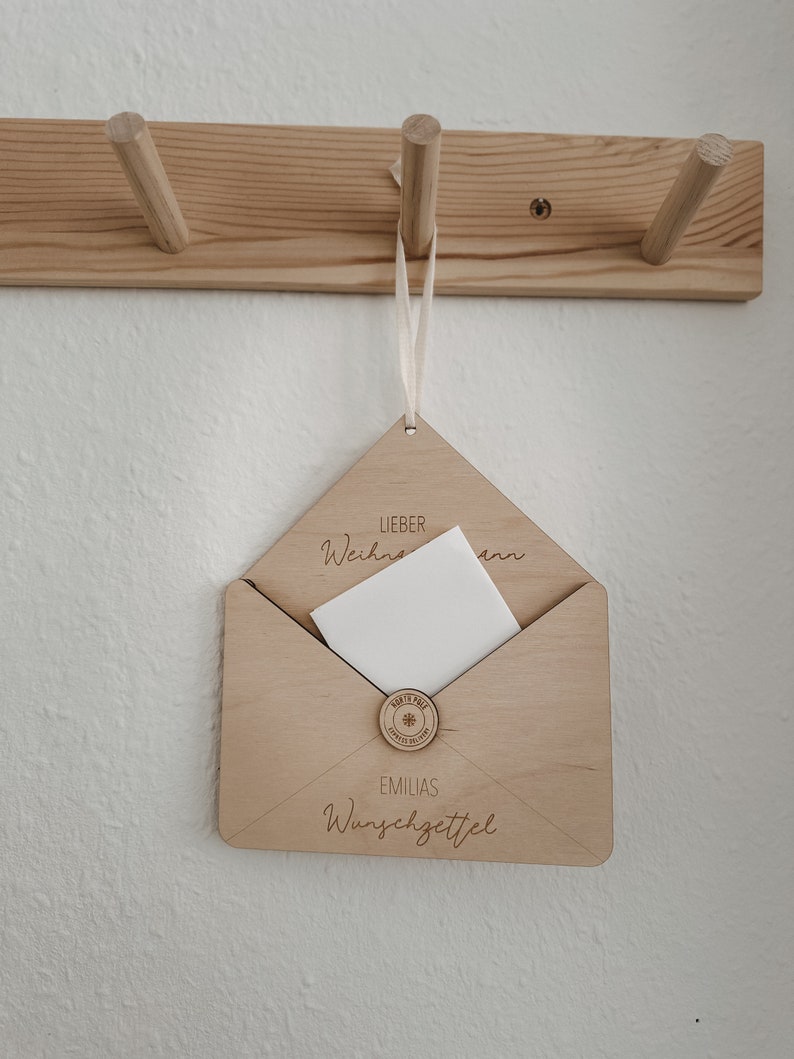 May include: A wooden envelope with a white card inside. The envelope has the text "Lieber Weihnachtsmann" and "Emilias Wunschzettel" engraved on it.