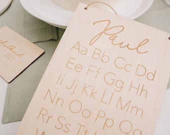 May include: A wooden sign with the name "Paul" written in cursive at the top. The sign features the alphabet in lowercase letters, with each letter repeated twice. The sign is light brown and has a slightly distressed look.