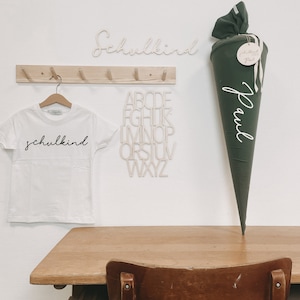 Könnte beinhalten: Ein weißes T-Shirt mit dem Wort "schulkind" in schwarzer Schrift. Ein Holzschild mit dem Wort "schulkind" in Schrift. Ein grüner Schultüte mit dem Namen "Paul" in weißer Schrift. Ein Holzschild mit dem Alphabet darauf.