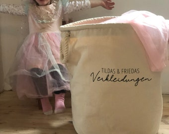 May include: A young girl wearing a pink unicorn costume stands next to a large white canvas storage bin with the text "Tildas & Friedas Verkleidungen" printed on it.