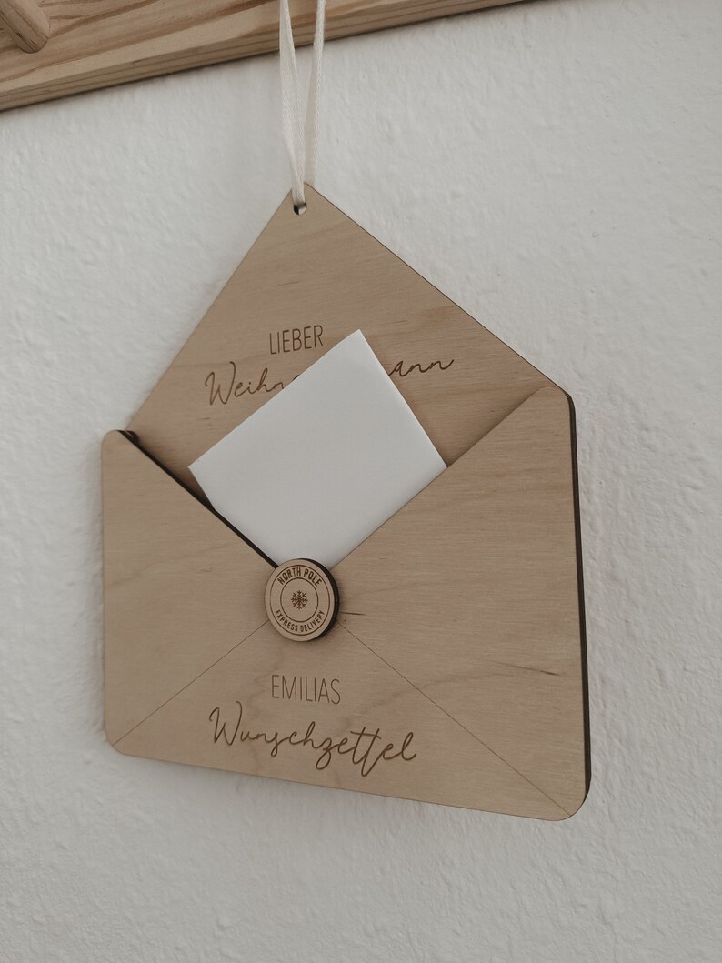 May include: A wooden envelope with a white card inside. The envelope has the text "Lieber Weihnachtsmann" and "EMILIAS Wunschzettel" on it. There is a round wooden seal with the text "North Pole Express Delivery" and a snowflake.