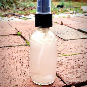 May include: A clear glass bottle with a black spray top filled with a light pink liquid. The bottle is sitting on a brick surface.