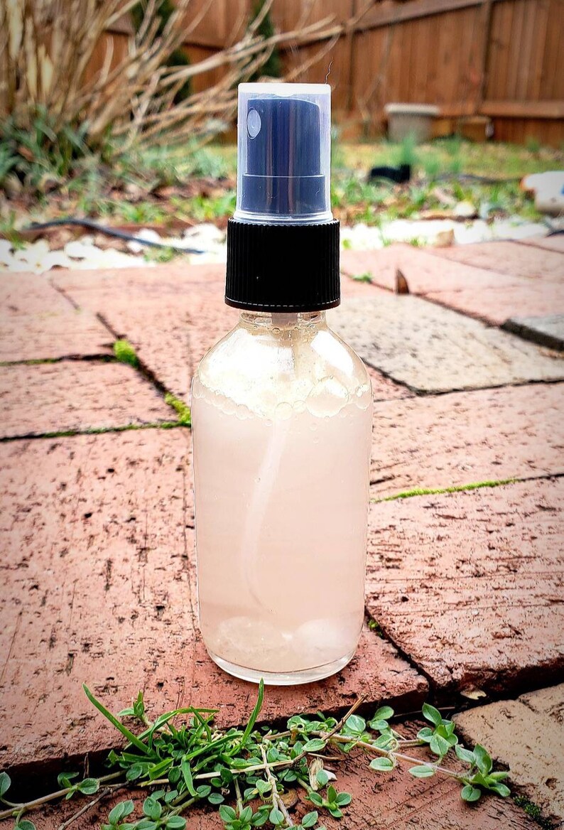 May include: A clear glass bottle with a black spray top filled with a light pink liquid. The bottle is sitting on a brick surface.