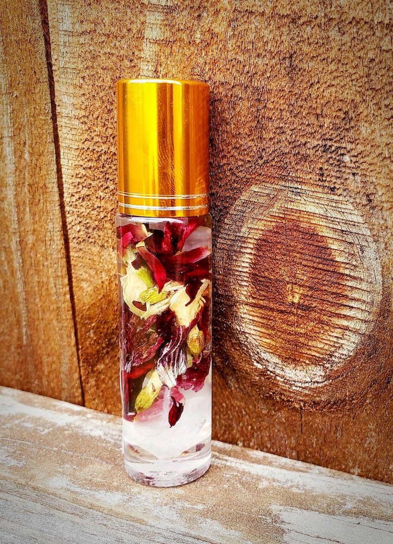 May include: A clear glass roller bottle with a gold lid filled with dried red and green flower petals and a white crystal at the bottom.