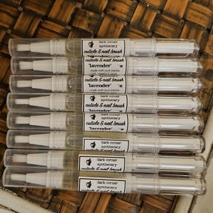 May include: A stack of ten clear plastic tubes with white caps and a white brush applicator. Each tube has a black and white label that reads "dark corner apothecary cuticle & nail brush 'lavender'".