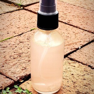 May include: A clear glass bottle with a black spray top filled with a light pink liquid. The bottle is sitting on a brown brick surface.