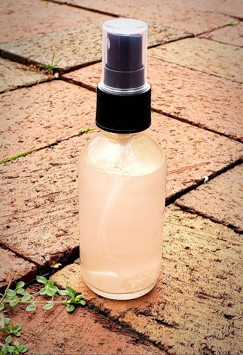 May include: A clear glass bottle with a black spray top filled with a light pink liquid. The bottle is sitting on a brown brick surface.