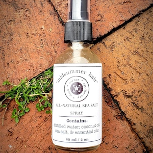 May include: A clear glass bottle with a black spray top and a white label that reads 'midsummer hair' corner apothecary. The label also says 'All-Natural Sea Salt Spray' and 'Contains: distilled water, coconut oil, sea salt, & essential oils' and '60 ml / 2 oz.'