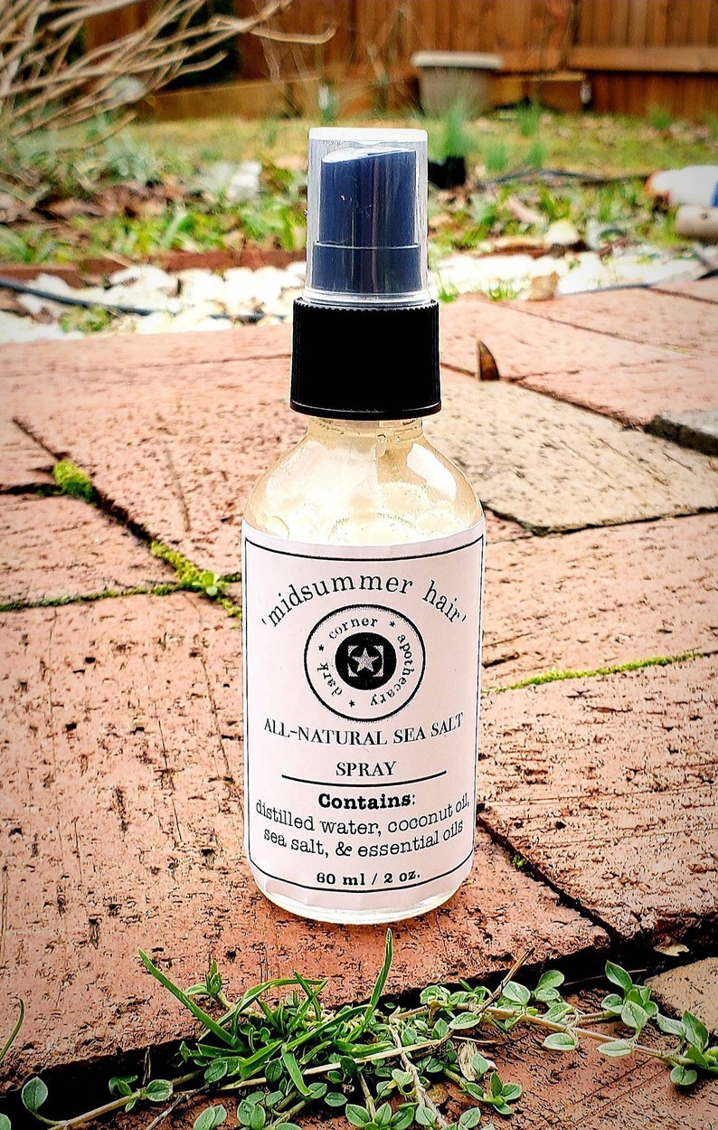 May include: A clear glass bottle with a black spray top and a white label that reads "midsummer hair corner apothecary" and "ALL-NATURAL SEA SALT SPRAY". The label also lists the ingredients: "distilled water, coconut oil, sea salt, & essential oils" and the size: "60 ml / 2 oz."