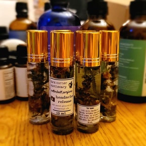 Puede incluir: Cuatro botellas de vidrio ámbar de 10 ml con tapas doradas llenas de un líquido transparente y hierbas secas. La etiqueta en la botella del centro dice "dark corner apothecary rollerball perfume headache release with sweet almond oil & essential oils 10 ml."