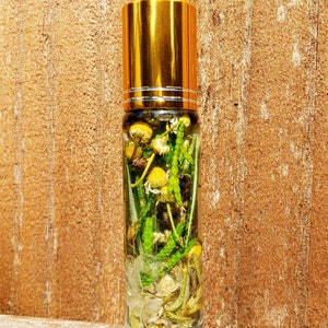 May include: A clear glass roller bottle with a gold lid filled with dried flowers and herbs. The bottle is sitting on a wooden surface.
