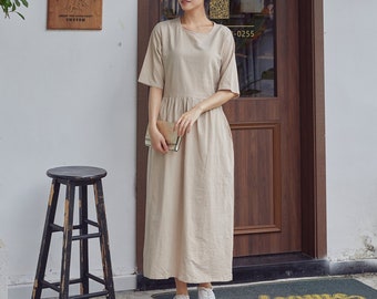 May include: A woman wearing a beige linen dress with a round neckline and 3/4 sleeves. She is standing in front of a wooden door with a sign that says "Opening Time Tuesday-Sunday 10:30-19:30".