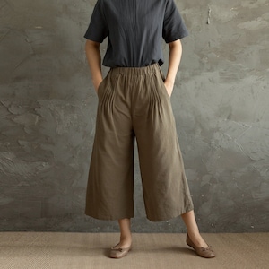 May include: A woman wearing a dark gray short-sleeved shirt and brown wide-leg cropped pants. She is standing in front of a gray textured wall.