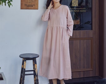 May include: A pale pink, long-sleeved, linen dress with a gathered waist and a round neckline. The dress has a relaxed fit and is shown on a model standing in front of a wooden door with a sign that says "Tuesday-Sunday, Open 10:30, Close 19:30, 33-9253-0255".