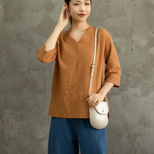 May include: A woman wearing a brown, three-quarter sleeve shirt with a white crossbody bag. She is standing in front of a gray textured wall.